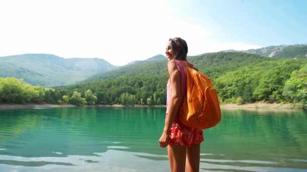 Vista trasera Turista mujer de pie levantando las manos sobre el fondo del lago de montaña y colinas . — Vídeo de stock