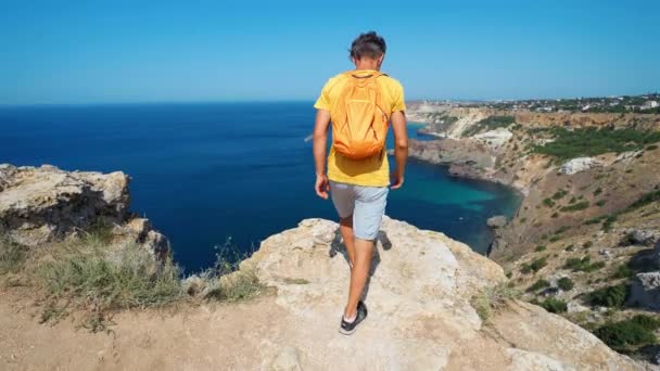 Joven viajero en camiseta naranja y pequeña mochila que llega al borde del acantilado con hermosa vista al mar — Vídeos de Stock