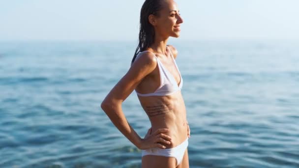 Slim femme avec un corps parfait en bikini blanc debout confiant sur fond de mer et bain de soleil . — Video