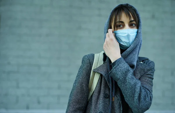 Concept coronavirus pandemic, quarantine, isolation period. woman in hood wearing face mask for protection of coronavirus, standing against grey wall in city street,