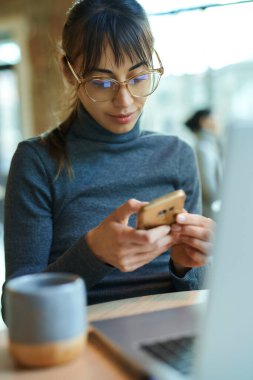 Akıllı telefondan bildiri okuyan konsantre genç bir kadın kafede dizüstü bilgisayar başında oturuyor. Kadın serbest çalışan cep telefonuyla internet bankacılığı uygulaması üzerinden para gönderiyor.