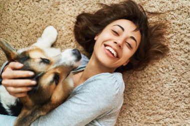 Güzel bir evcil hayvanla halıya uzanmış gülümseyen kız portresi. Galli Corgi köpeğini kucaklayan ve okşayan genç bir kadın. Evde kalmak, köpekle arkadaşlık, mutlu anlar..