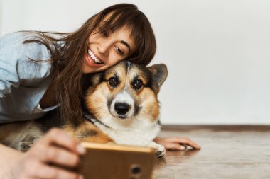 Genç bir kadının portresi gülüyor tatlı bir Galli Corgi köpeğine sarılıyor ve akıllı telefon kamerasında evcil hayvanıyla selfie çekiyor. Evde kalmak, köpekle arkadaşlık etmek, fotoğraf çekmek..