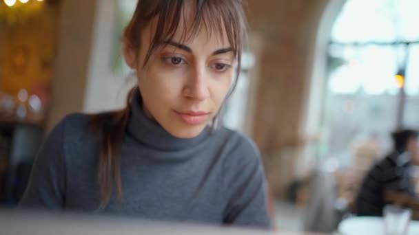 Νεαρή γυναίκα που επικεντρώνεται στην οθόνη και δακτυλογράφηση σε φορητό υπολογιστή, ενώ κάθεται στο γραφείο στο χώρο εργασίας ή καφέ. — Αρχείο Βίντεο