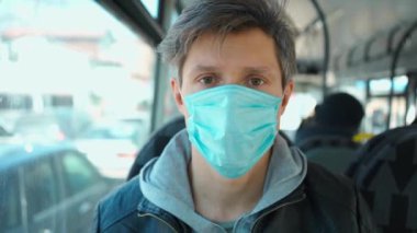 Korumacı tıbbi maskeli genç Avrupalı adam şehir ulaşımında otururken doğrudan kameraya bakıyor.