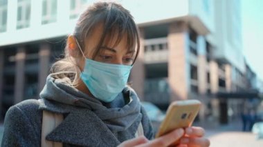 Yavaş çekim kameralı koruyucu maskeli kadın dışarıda akıllı telefon kullanıyor. Kadın cep telefonuna bakıyor, sosyal medyada iletişim kuruyor.