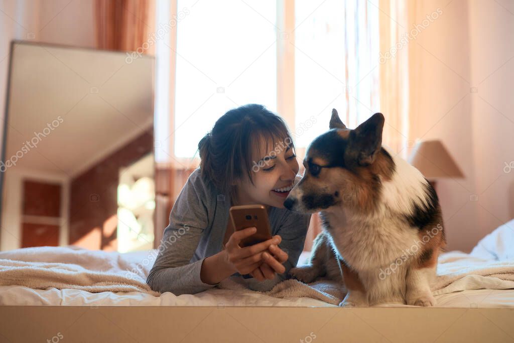Smiling young woman lying on bed in bedroom with her cute Welsh Corgi dog, showing something on smartphone. Concept stay at home, friendship with pet, spending time together.