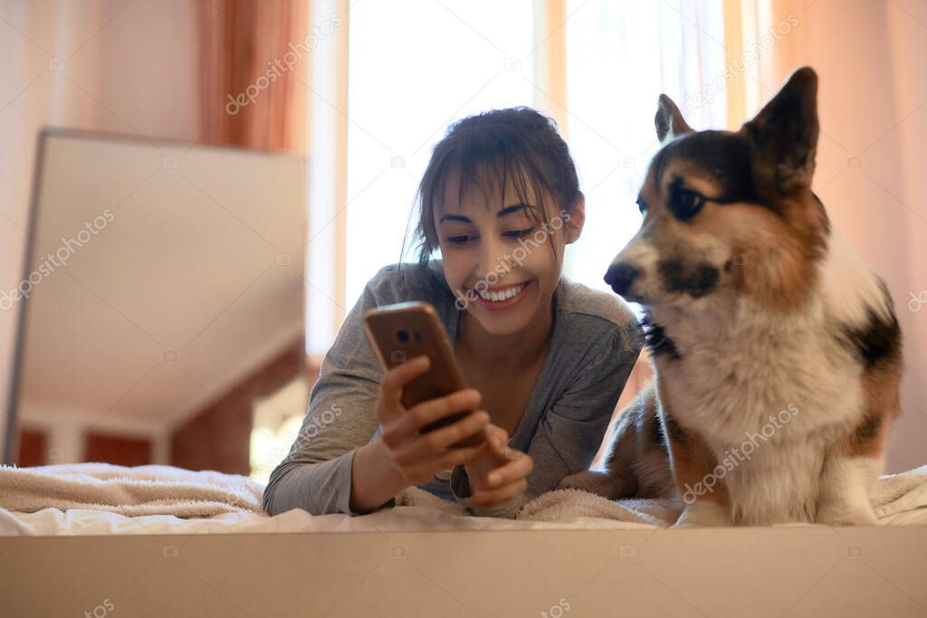 Beautiful female owner lying on bed in bedroom with her cute Welsh Corgi dog, holding smartphone, watching video online together. Concept stay at home, friendship with pet, spending time together.