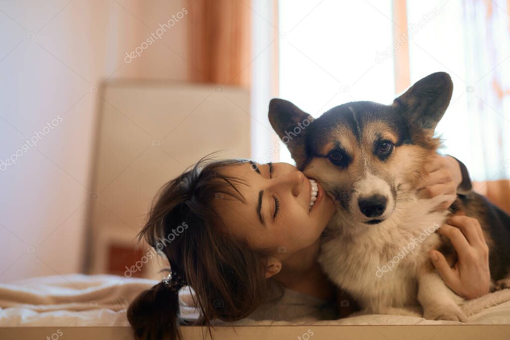 Happy woman with dog in bed at home, hugging funny adorable dog Welsh Corgi with eyes closed. Girl enjoying good day and posing with pet. Domestic cozy atmosphere.