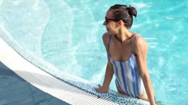 Beautiful sexy smiling woman in swimsuit and sunglasses standing at pool edge in water — Stock Video