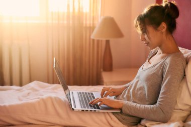 Yatakta otururken dizüstü bilgisayarda yazan başarılı kadın serbest yazar. Pijamalı esmer kız evden çalışıyor, uzaktan çalışıyor, internet işinde çalışıyor, arka planda pencereden gün ışığı görüyor.