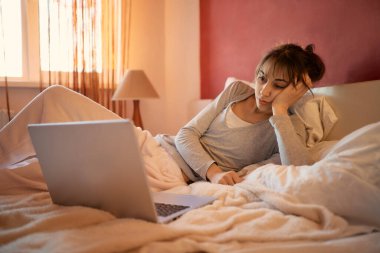 Evde pijamalarıyla yalnız başına takılan, yatakta yatıp dizüstü bilgisayar izleyen, güneş ışığında internette gezinen bir kadın. Evde kalıp konsepti karantinaya almak.