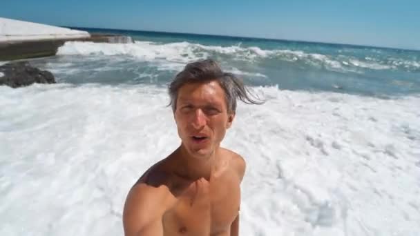 Щасливий сміх засмучений спортивний молодий чоловік бере відео селфі під час прогулянки вздовж пляжу з хвилястим морем — стокове відео
