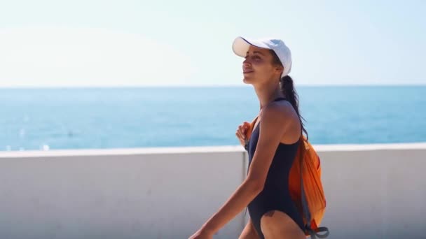 Стильная сексуальная дама в черном купальнике, белой шапке и оранжевом рюкзаке ходит по бухте у причала — стоковое видео