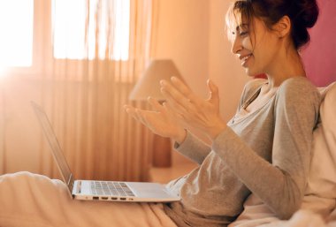 Neşeli bir kadın evde arkadaşıyla dizüstü bilgisayar ve video görüşmesi yapıyor. Yatak odasında güneş ışığı var. Kablosuz internet kullanarak sosyal ağlarda dizüstü bilgisayarda sohbet eden olumlu kız