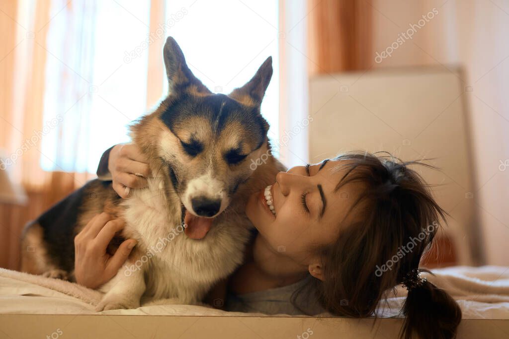 Smiling woman with dog in bed at home, hugging funny yawning dog Welsh Corgi Girl enjoying good day and posing with pet. Domestic cozy atmosphere.