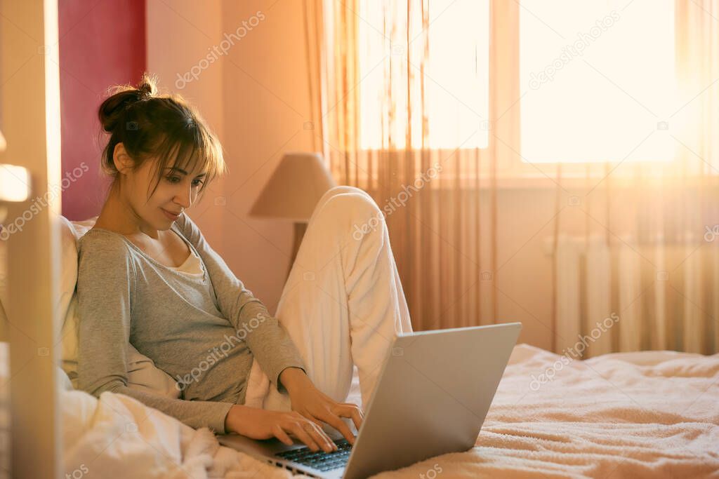 Concentrated young woman relaxing in bed at home, using laptop computer, sunlight in bedroom. Concept remote work, internet shopping, checking social network, reading news or communicating online