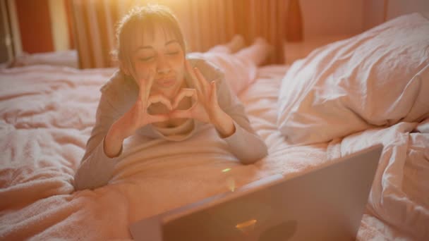 İnternette görüntülü konuşma yapan mutlu bir kadın, dizüstü bilgisayar kullanıyor, erkek arkadaşına hava öpücüğü gönderiyor evde yatarken. — Stok video