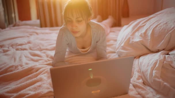 Jovem mulher de pijama usando computador portátil enquanto relaxa na cama em casa, luz solar no quarto. — Vídeo de Stock