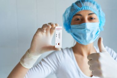 Koruyucu yüz maskesi takmış doktor ya da hemşire. Elleriyle olumlu jest yapıyor. Başparmakları havada, ellerinde COVID-19 testi var..