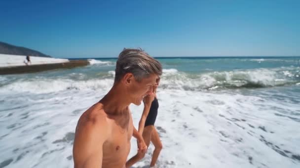 Glücklich gebräunter Mann und Frau in Badebekleidung joggt freudig am welligen Meeresstrand — Stockvideo