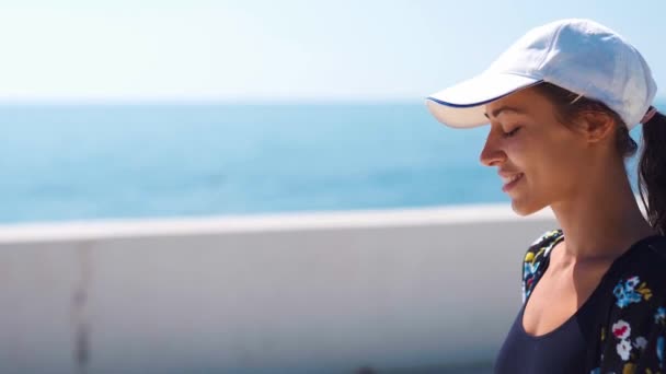 Retrato de primer plano exterior Hermosa mujer deportiva con estilo en traje de baño negro y gorra blanca caminando a lo largo de la bahía junto al mar — Vídeo de stock