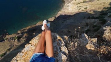 Uçurumun kenarında oturan sarı elbiseli bronzlaşmış kadın bacakları ve deniz manzarası inanılmaz..