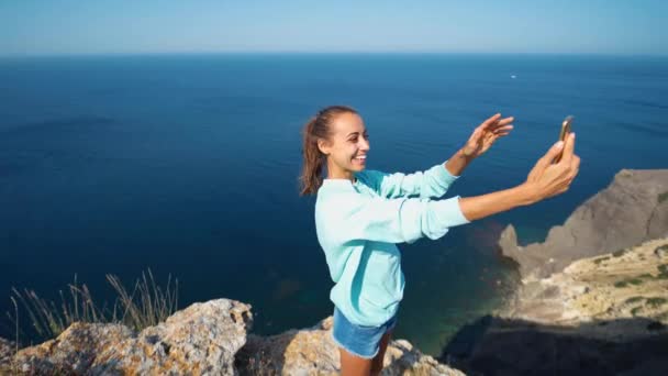 Jonge vrouwelijke reiziger maken selfie op klif rand met prachtig uitzicht op zee, lachen en glimlachen naar de camera, het maken van grappige gezicht. — Stockvideo