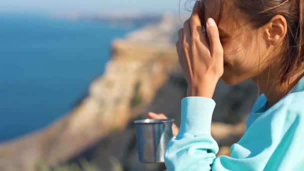 Młoda kobieta siedzi na krawędzi urwiska, pije kawę lub herbatę, ciesząc się niesamowitym widokiem na przylądek i zatokę morską. — Wideo stockowe