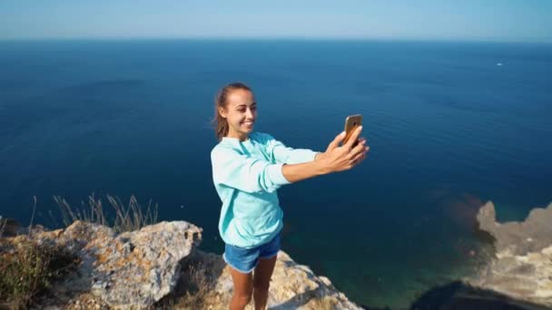 Joven viajera haciendo selfie en el borde del acantilado con hermosa vista al mar, riendo y sonriendo a la cámara, haciendo cara divertida . — Vídeos de Stock