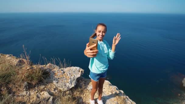 Весёлая улыбающаяся девушка с видеочатом на высоком утёсе над морем . — стоковое видео