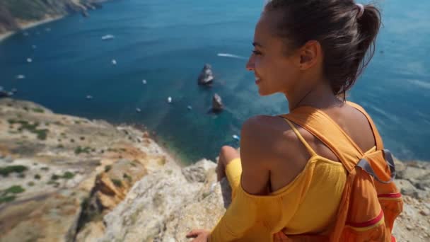 Chica sonriente en vestido amarillo y zapatillas blancas sentadas en el borde del acantilado sobre la costa — Vídeos de Stock