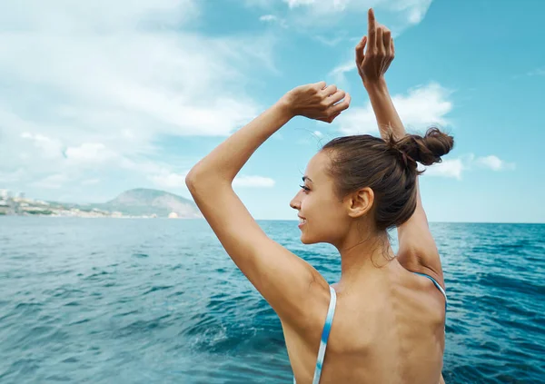 Huoleton hymyilevä rusketus nainen uimapuvussa nauttii merinäköalasta ja poseeraa kädet sinistä taivasta vasten . — kuvapankkivalokuva