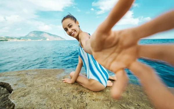 Mooie bruine vrouw in badpak zitten op stenen strand met de hand reiken, volg me tegen uitzicht op zee. — Stockfoto