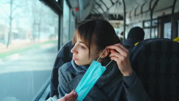 Молодая брюнетка в сером пальто надевает защитную медицинскую маску в городском общественном транспорте — стоковое видео