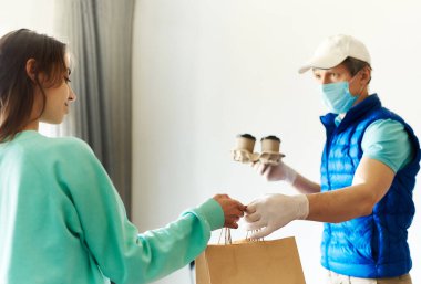 Kadın teslimatçıdan paket alıyor. Üniformalı kurye maske eldivenleri, yiyecek ve poşetler için kağıt kutular tutuyor..
