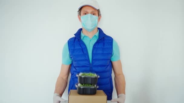 Entrega durante la cuarentena. Mensajero en máscara protectora y guantes sobre fondo blanco sosteniendo paquetes de papel y contenedores para comida para llevar — Vídeos de Stock