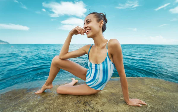 Vakker, smilende kvinne i badedrakt som sitter på steinstranden og nyter havutsikt og blå himmel . – stockfoto