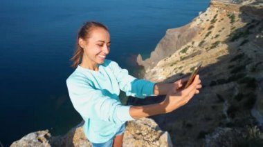 Genç bayan gezgin uçurumun kenarında güzel deniz manzaralı selfie yapıyor, kameraya gülüyor ve gülüyor, komik bir yüz ifadesi takınıyor..