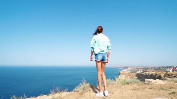 Vista posterior de la joven despreocupada de pie con las manos levantadas en el borde del acantilado — Vídeo de stock