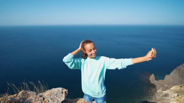 Felice ragazza sorridente in felpa blu brillante e pantaloncini jeans facendo selfie sul bordo della scogliera — Video Stock