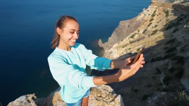 Młoda kobieta podróżnik co selfie na krawędzi klifu z pięknym widokiem na morze, śmiech i uśmiech do kamery, co zabawne twarz. — Wideo stockowe