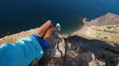 Uçurumun kenarında oturan cesur genç kadın kameraya gülümseyerek el sallıyor.