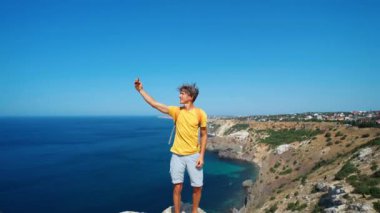 Genç erkek turist, uçurumun kenarında özçekim yapıyor ve gülümsüyor. Güzel deniz manzarası ve harika doğanın tadını çıkarıyor..