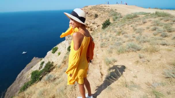 Feliz mujer sonriente en vestido amarillo brillante y sombrero con mochila de pie en el borde del acantilado — Vídeos de Stock