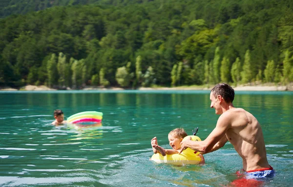 Atlético homem feliz se divertindo com crianças no lago da montanha com água azul clara. Natação familiar, banho quente de verão — Fotografia de Stock