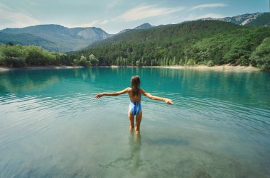 Mayo giymiş özgürlükçü kadın dağ gölünde kolları açık ve arka planda dağlarla ayakta duruyor.