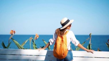 Yavaş çekim genç bayan turist hasır şapka ve turuncu sırt çantası güneşli bir günde deniz manzarasının tadını çıkarıyor..