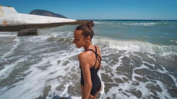 Alegre joven en traje de baño negro y gafas de sol de pie en el agua en la orilla del mar — Vídeos de Stock