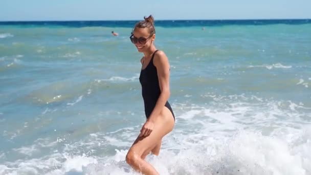 Χαρούμενη νεαρή γυναίκα με μαύρο μαγιό και γυαλιά ηλίου στέκεται στο νερό στην παραλία — Αρχείο Βίντεο